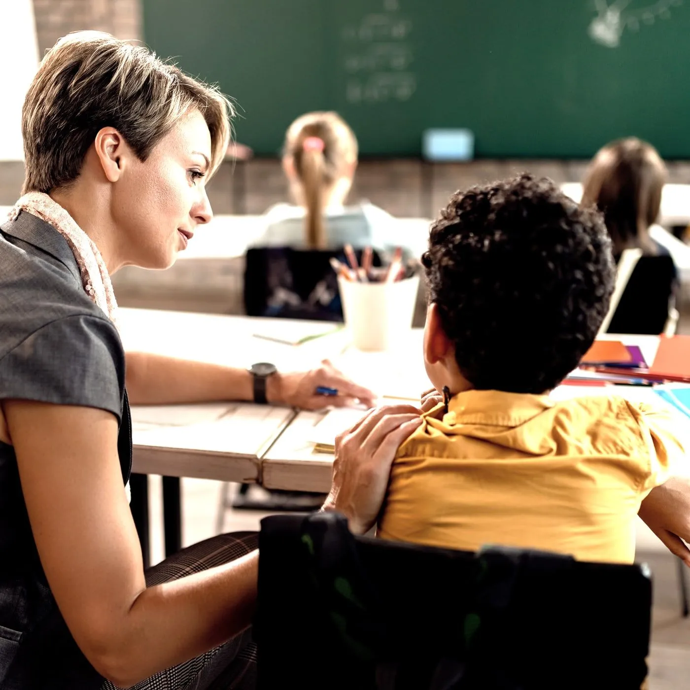 Eine Schulbegleiterin sitzt neben einem Jungen im Klassenzimmer und zeigt ihm etwas in seinem Heft
