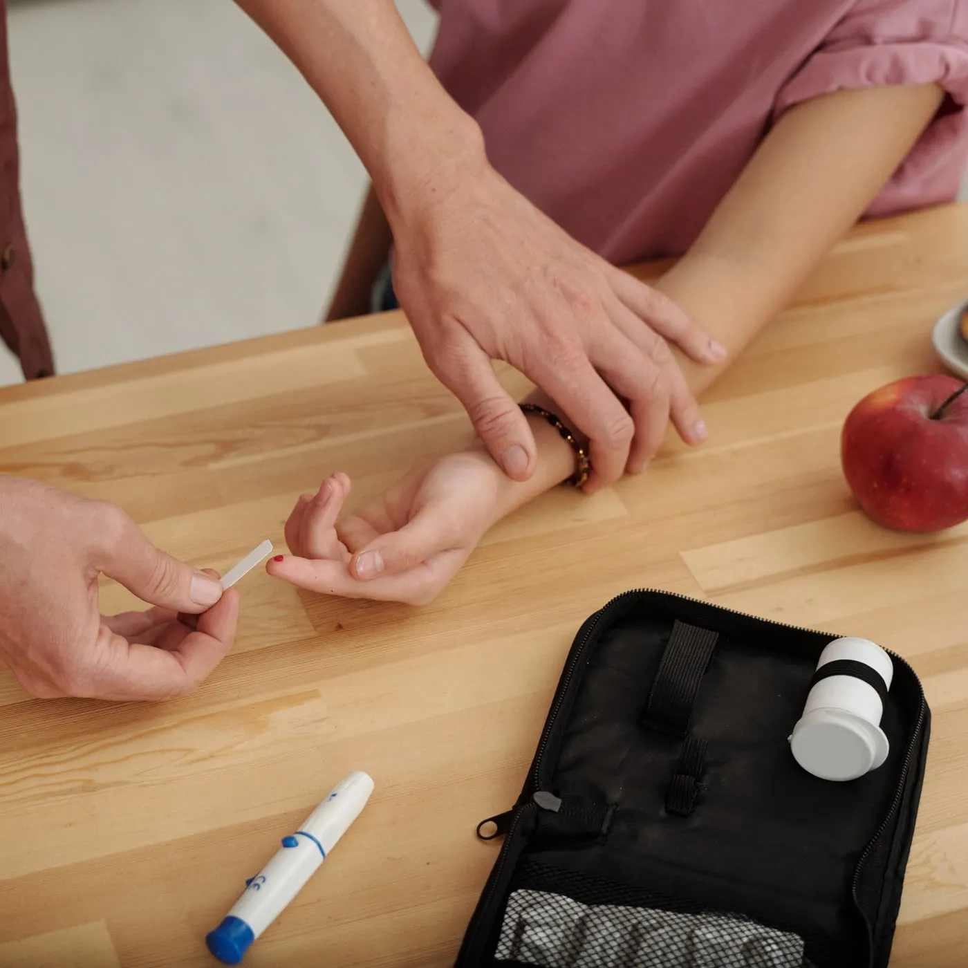 Eine erwachsene Hand nimmt am Fingerkuppen eines Kindes Blut für die Blutzuckermessung ab, daneben liegen Messgerät und Diabetestasche